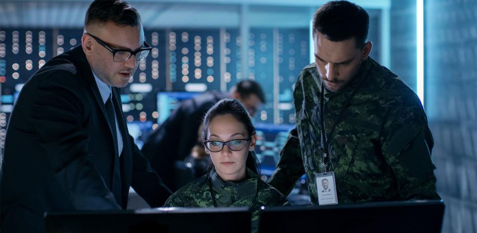 A man and woman in military fatigues accompanied by a male government official looking at a computer in a control room.