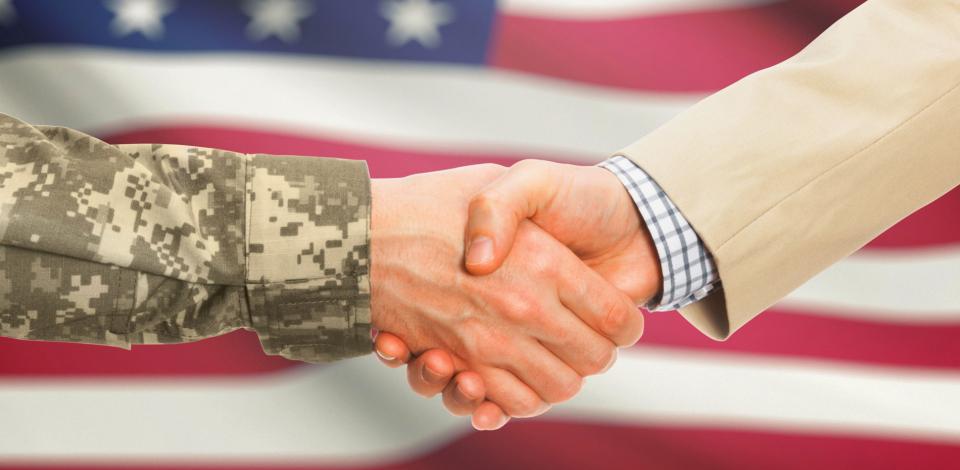 Two hands shaking, one in suit and one in military uniform with the american flag as a backdrop