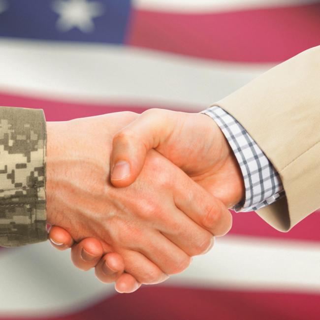 Two hands shaking, one in suit and one in military uniform with the american flag as a backdrop